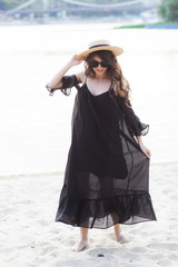 Fototapeta na wymiar Beautiful young brunette woman in black dress, black sunglasses and straw hat walking on the sand on the beach and posing at sunrise in summer