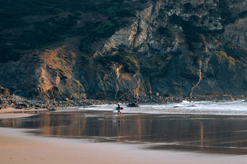 Very wide and open beach with tide line and mountain rock, surfer walks towards waves with his surfing board, ready to take on adventures