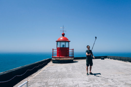 Handsome young man in traveler outfit, stands near bright red lighthouse, sets up timer on smartphone on selfie stick, ready to make photo for social media
