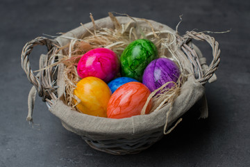 Multi-colored painted Easter eggs in a basket with straws on a dark stone background. Happy Easter holiday card or banner. 