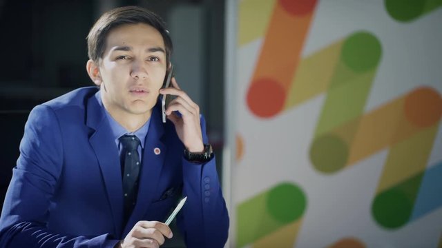 A man in a business suit is talking on the phone reluctantly. A man in blue attire sits at a table and communicates with a partner about an unpleasant problem. The male figure holds a pencil in his