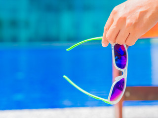 Woman hand holding sunglasses and lying on deckchair by the swimming pool. Vacation and relaxation, summer travel concept.