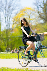 Portrait view of girl on bicycle wearing on black short dress. Young happy Woman riding along road on green spring  outdoor Park. Sporty young girl riding a bicycle on a sunny morning, view from face