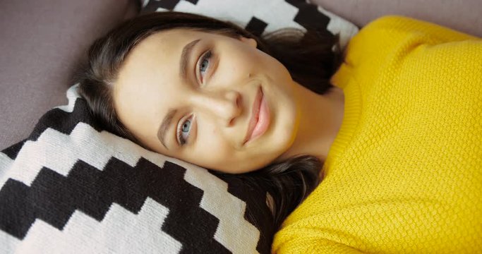 Portrait of attractive woman smiling to the camera. Portrait of beautiful young woman in yellow sweater lying in the pillow on the couch in the living room. Close up shot