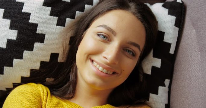 Portrait of pretty woman smiling to the camera. Portrait of beautiful young woman in yellow sweater lying in the pillow on the couch in the living room. Close up shot