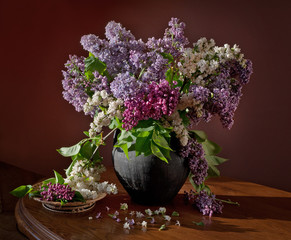 Still life with blooming branches of lilac and tulips in a clay jug on a wooden table.