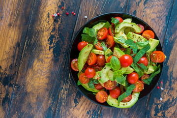 Vegetarian salad with avocado, cherry tomatoes and basil leaves on a black plate on the wooden rustic table, top view. - 164016418