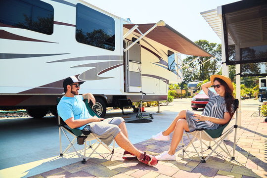 Young couple sits near camping trailer,smiling.Men talks on mobile phone and uses electronic device, woman relax on chair near car and palms.Family spending time together on vacation in rv park