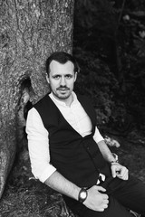 the groom in a white shirt and a black vest sits under a tree