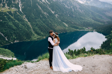 Fototapeta na wymiar bride with the beautiful blue dress and groom hugging with views of the beautiful green mountains and lake with blue water
