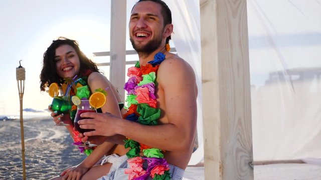 laughing couple from Hawaii lei on neck on Tropical Islands, guy and girl to exotic vacation happy friends drink cocktail in ocean coast, young people in bathing suits in sunlight by ocean beach party