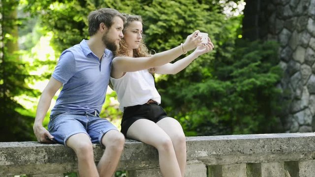 Couple with smartphone sitting on concrete wall taking selfie.