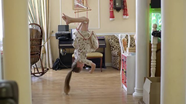 Wide shot of little girl in simple linen dress hanging upside down on rope ladder in living room