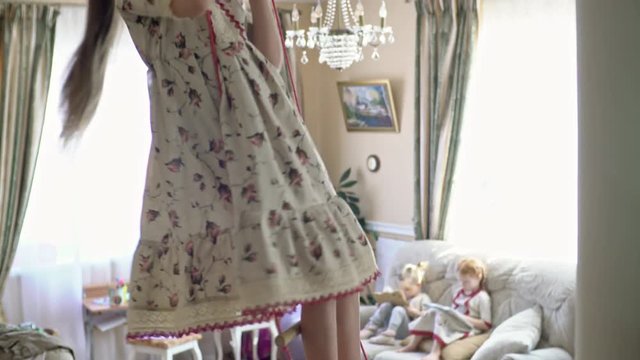 Tilt up of little girl in linen dress standing on trapeze hanging in playroom; her siblings sitting on sofa and playing on tablets in background