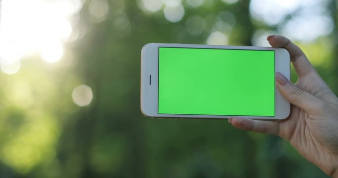 closeup young woman's hand holding keeps horizontally phone smartphone device green screen сhroma key, chromakey outdoor unfocused nature sunny day girl new technology development appliance