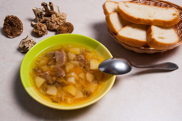 Mushroom soup in green plate with metal spoon, dried wild mushrooms and basket of bread on a light stone background.