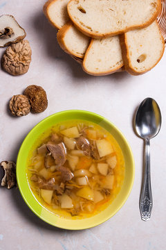 Mushroom soup in green plate with metal spoon, dried wild mushrooms and basket of bread on a light stone background.