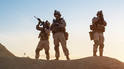 Squad of Three Fully Equipped and Armed Soldiers Standing on Hill in Desert Environment in Sunset Light.