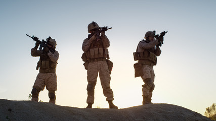 Squad of Three Fully Equipped and Armed Soldiers Standing on Hill in Desert Environment in Sunset...
