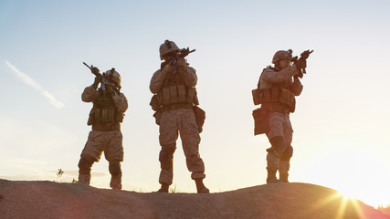 Squad of Three Fully Equipped and Armed Soldiers Standing on Hill in Desert Environment in Sunset Light.