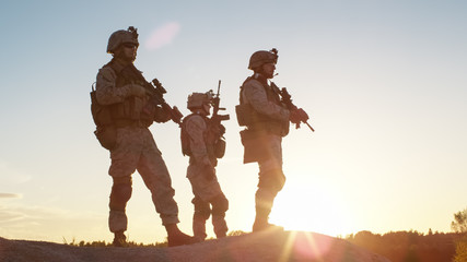 Squad of Three Fully Equipped and Armed Soldiers Standing on Hill in Desert Environment in Sunset...