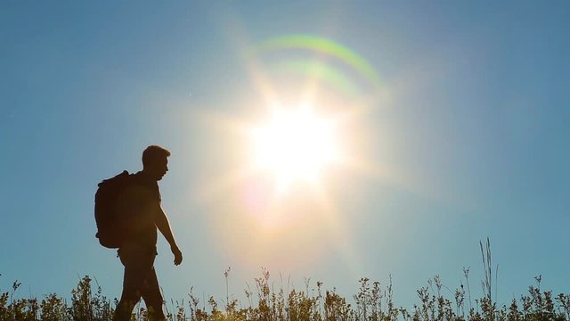 Adult man on summer vacations in countryside. Male silhouette of tired guy hiking. Person walks along grassy hill at sunset sunlight and blue sky background, resting, wiping sweat.