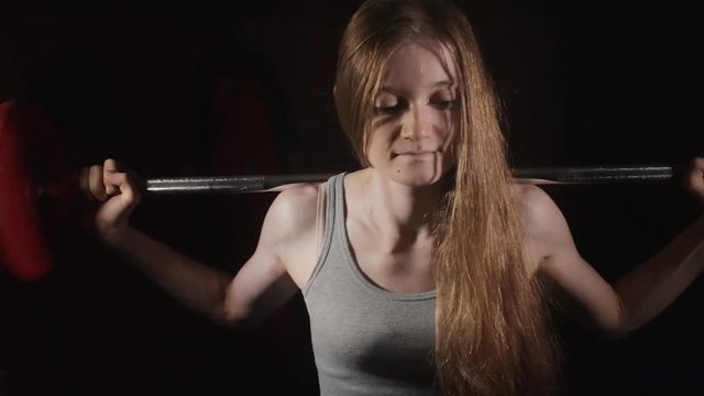 Young woman doing exercise with a barbel in the dark gym at the black background. Resting after exercises with a barbell.
