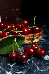Several red sweet cherries and big green leaf on the table. Fresh organic cherry in yellow wooden basket on dark marble background..