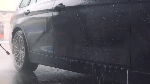 A man washes his dark gray car with a hose in a self-service car wash station - closeup on the bottom