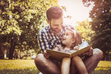 Single father sitting on grass with little daughter and reading book story. Little girl sitting on father lap.