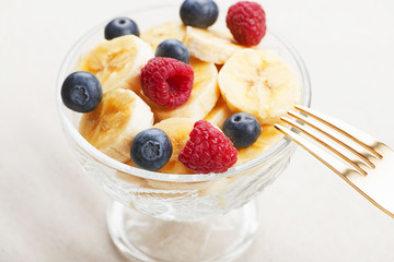 Tasty breakfast of bowl with banana, blueberries and raspberries next to fork on tablecloth.