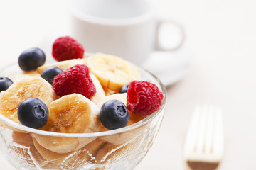 Tasty breakfast of bowl with banana, blueberries and raspberries next to cup of coffee.