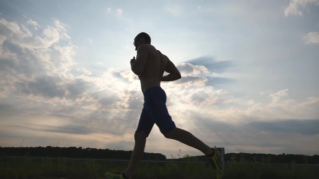 Silhouette of muscular man jogging in the country road at sunset. Male jogger training for marathon run outdoor. Athlete exercising and running against blue sky. Sport and active lifestyle Slow motion