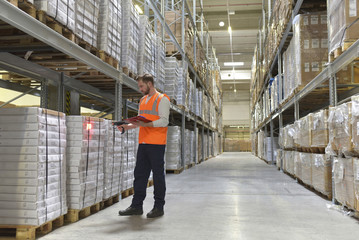 Man in factory hall wearing safety vest holding clipboard scanning goods
