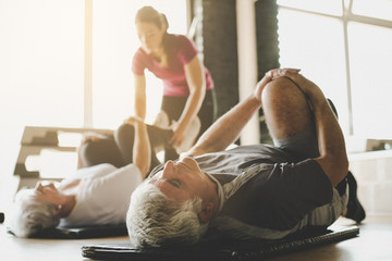 Senior couple workout in rehabilitation center. Personal trainer helps elderly couple to do stretching on the floor. Focus on man. - 164050483