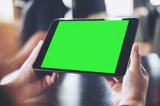Mockup image of woman's hand holding black tablet pc with blank green screen in cafe with blur background