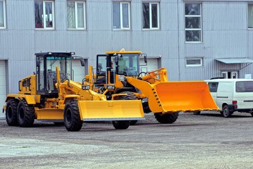 Два современных оранжевых трактора с ковшом возле стены дома