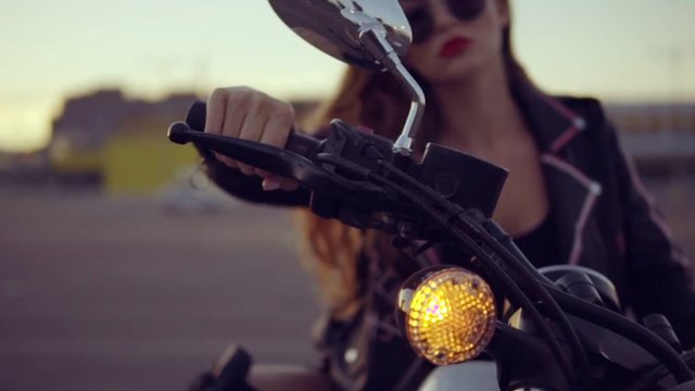 Portrait of a female biker in leather jacket and shorts sitting on her bike and holding a handlebar. Close up of young sexy curly woman in sunglasses on the chopper