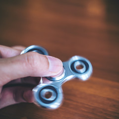 A man's hand holding and playing metal silver color fidget spinner with wooden table background