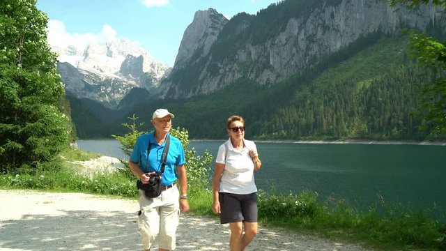 4k Travel Video, Happy Active Senior Couple On A Walk On Path Close To Mountain Lake, Man With Digital Camera
