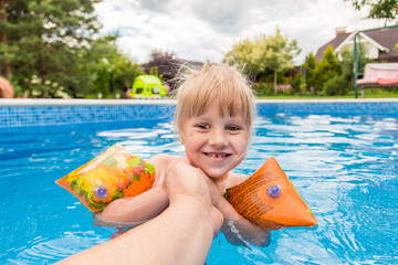 A cute blond baby girl swims at swimmig pool with blue colored water outdoors, smiles  and hold parent's hand.