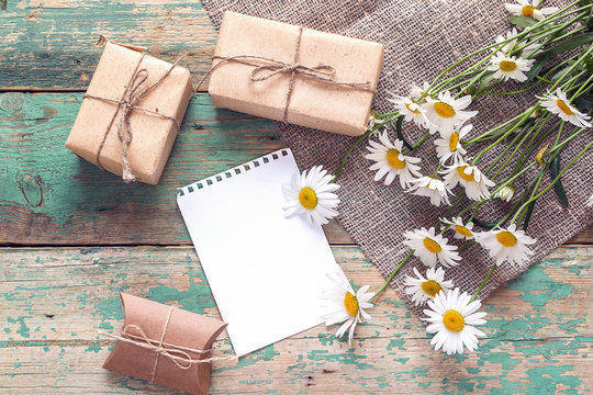 Bouquet of daisies with gift boxes and empty note pad for message on wooden background.