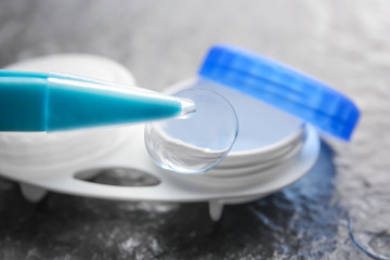 Tweezers with contact lens and blurred container on gray textured background