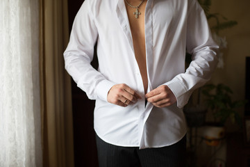 Groom buttons up white shirt standing before a window