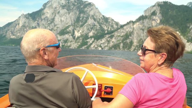 4k travel video, happy senior couple driving with small electric boat on austrian mountain lake on sunny summer vacation day
