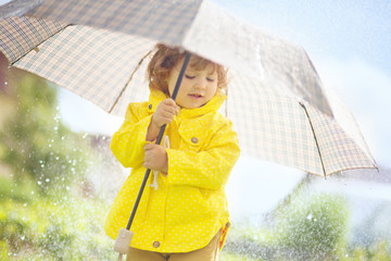 Cute toddler girl wearing yellow waterproof coat and boots with big adult umbrella having fun In...