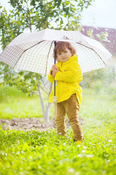 Adorable curly toddler girl wearing yellow waterproof coat holding big adult umbrella uder the pouring rain