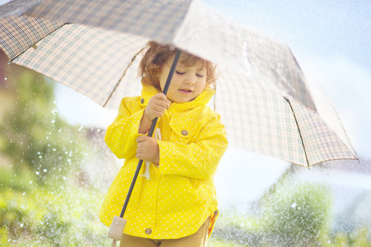 Cute toddler girl wearing yellow waterproof coat and boots with big adult umbrella having fun In the pouring rain. Warm and sunny autumn day