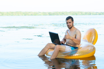 Businessman working on laptop sitting in inflatable ring in water and showing ok sign, free space.