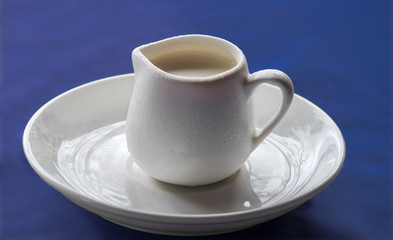 White ceramic creamer on a saucer with cold cream on a blue background.